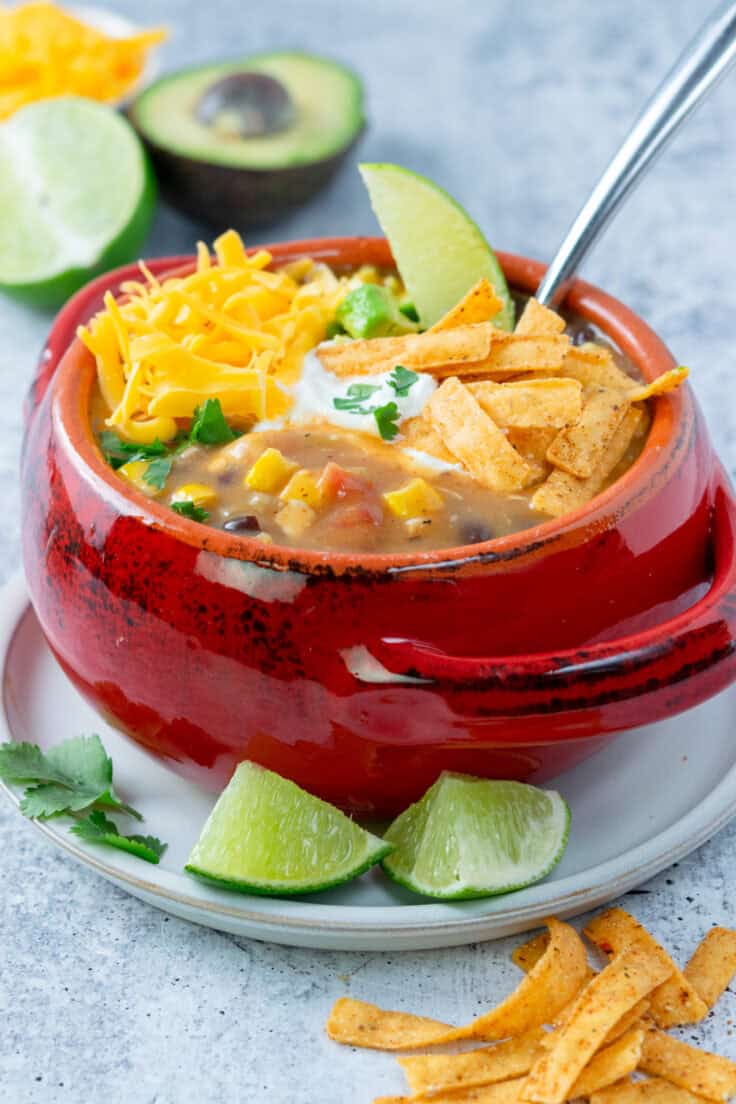 A red soup bowl on a plate that has two lime wedges and some cilantro. The bowl is full of a creamy chicken tortilla soup that's topped with cheddar cheese, sour cream, cilantro, tortilla strips and a lime wedge.