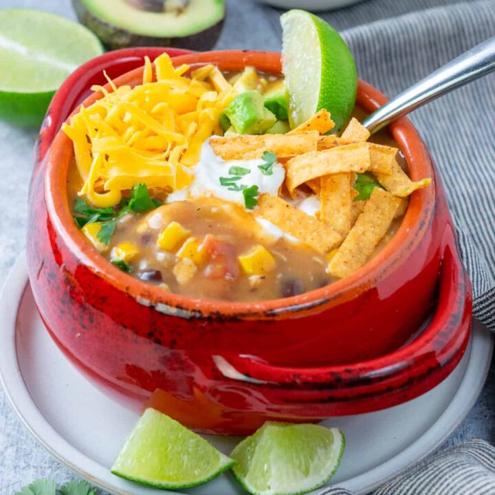 A bowl of homemade creamy chicken tortilla soup topped with cheddar cheese, sour cream, cilantro, tortilla chips and a lime wedge.