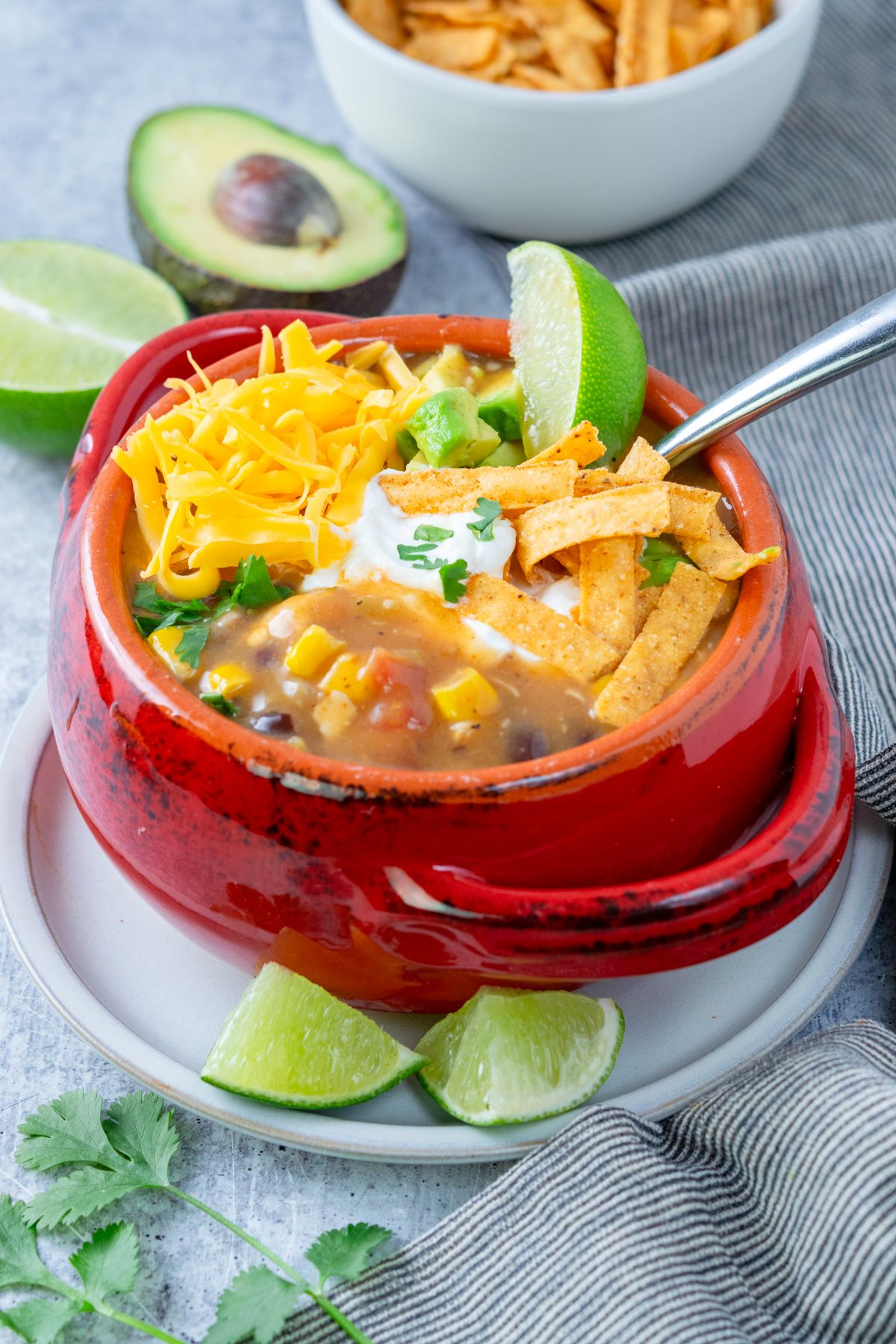 A bowl of homemade creamy chicken tortilla soup topped with cheddar cheese, sour cream, cilantro, tortilla chips and a lime wedge.