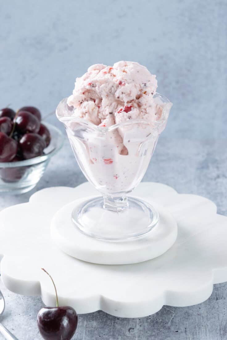 A big scoop of homemade Cherry Garcia ice cream in an ice cream glass that's sitting next to a bowl of cherries.