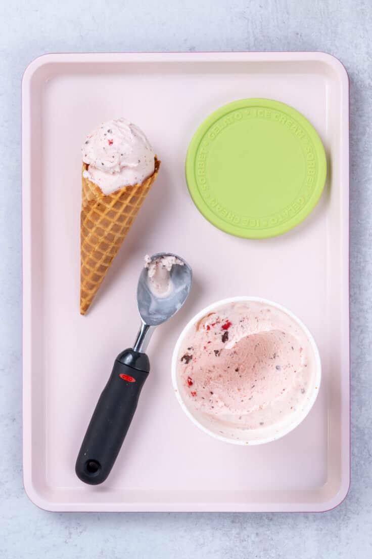 The ice cream container opened up and sitting on a pink tray with an ice cream scoop next to it, showing how the homemade Cherry Garcia firmed up after being placed in the freezer for a couple hours. There is also a waffle cone with two scoops of Cherry Garcia sitting on the pink tray along with the container's green lid.