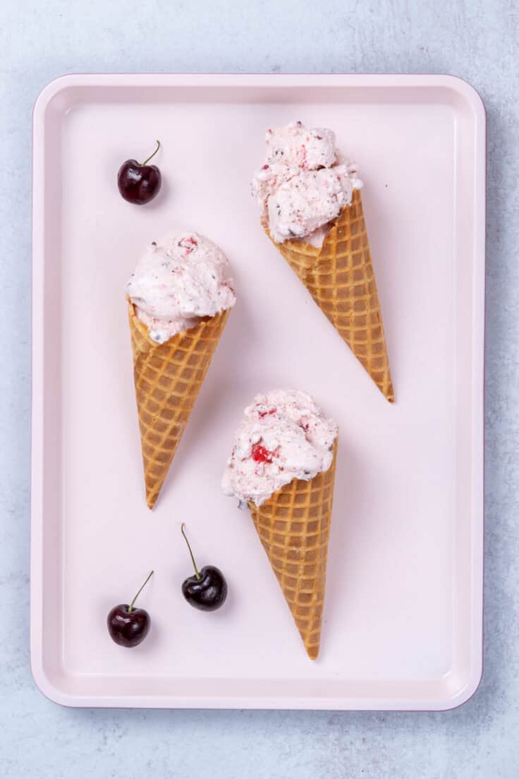 Three homemade, double-scoop Cherry Garcia waffle cones laying on a pink tray with three cherries with stems scattered around.