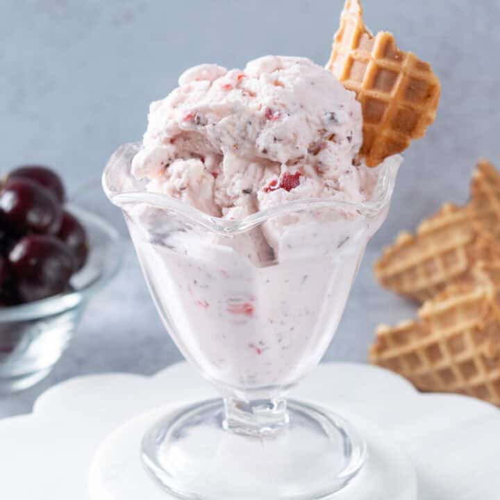 Homemade Cherry Garcia ice cream in an ice cream glass. The ice cream is garnished with a piece of waffle cone and the scoops of ice cream are next to two waffle cones and a bowl of cherries.