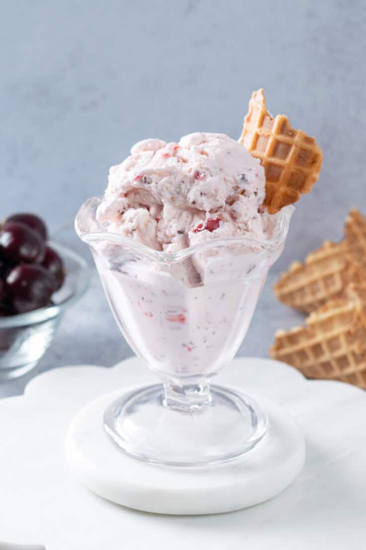 Homemade Cherry Garcia ice cream in an ice cream glass. The ice cream is garnished with a piece of waffle cone and the scoops of ice cream are next to two waffle cones and a bowl of cherries.