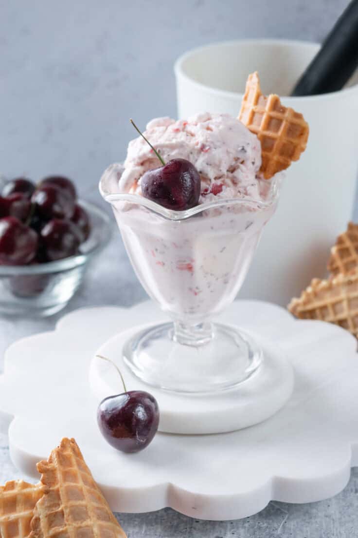 A big scoop of homemade cherry ice cream in an ice cream glass that's next to a bowl of cherries and some waffle cones and in front of a container with a scoop in it.