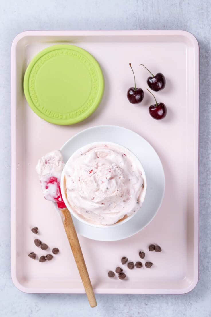 Putting the soft Cherry Garcia ice cream into a container with a red rubber spatula. The container is on a pink tray with some chocolate chips scattered around and three cherries with stems.