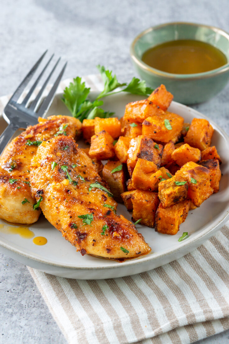 Two honey garlic chicken tenders dripping with sauce on a plate with roasted sweet potatoes. The meal is garnished with parsley and a fork is resting on the plate which is next to a small bowl of honey.