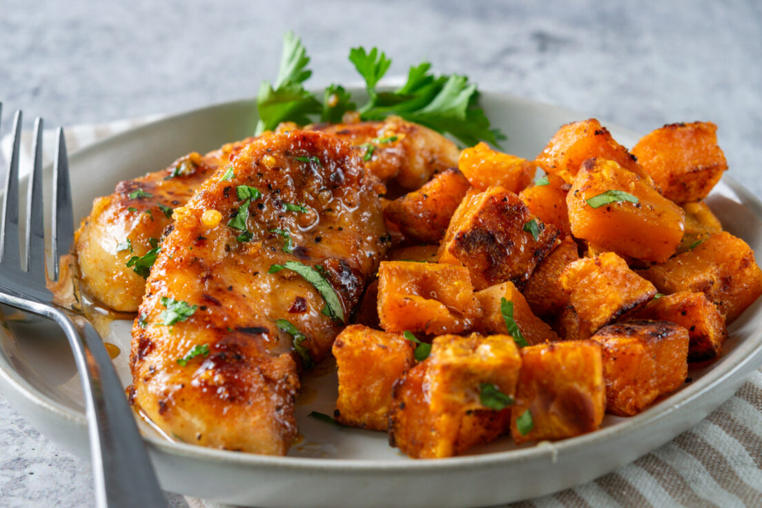 Juicy chicken tenders coated in honey and garlic next to a pile of roasted sweet potatoes on a plate with a fork on it.