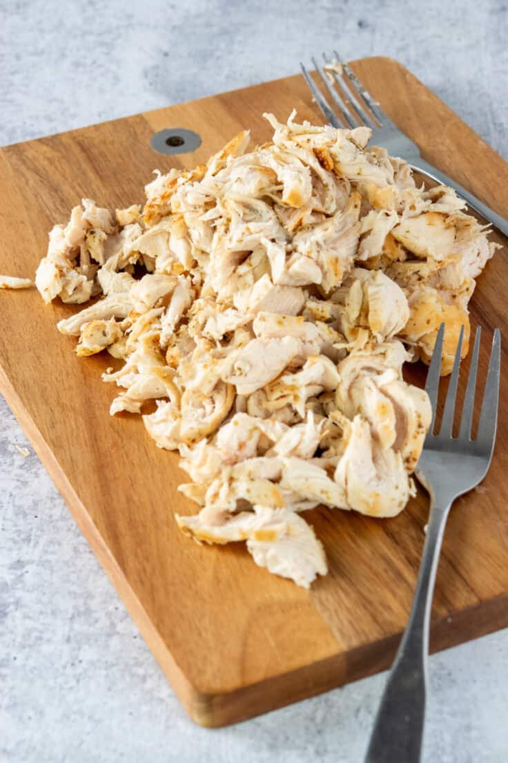 A wooden cutting board with a pile of shredded cooked chicken and two forks used that were used to shred the meat.