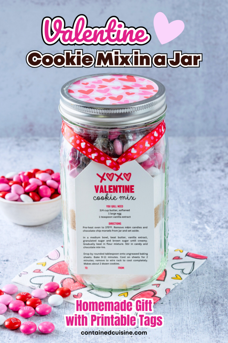 A mason jar filled with cookie mix that has a Valentine cookie mix gift tag on it. The cookie mix in a jar is decorated with valentine's day heart ribbon and the jar is next to a bowl full of Cupid M&M candies.