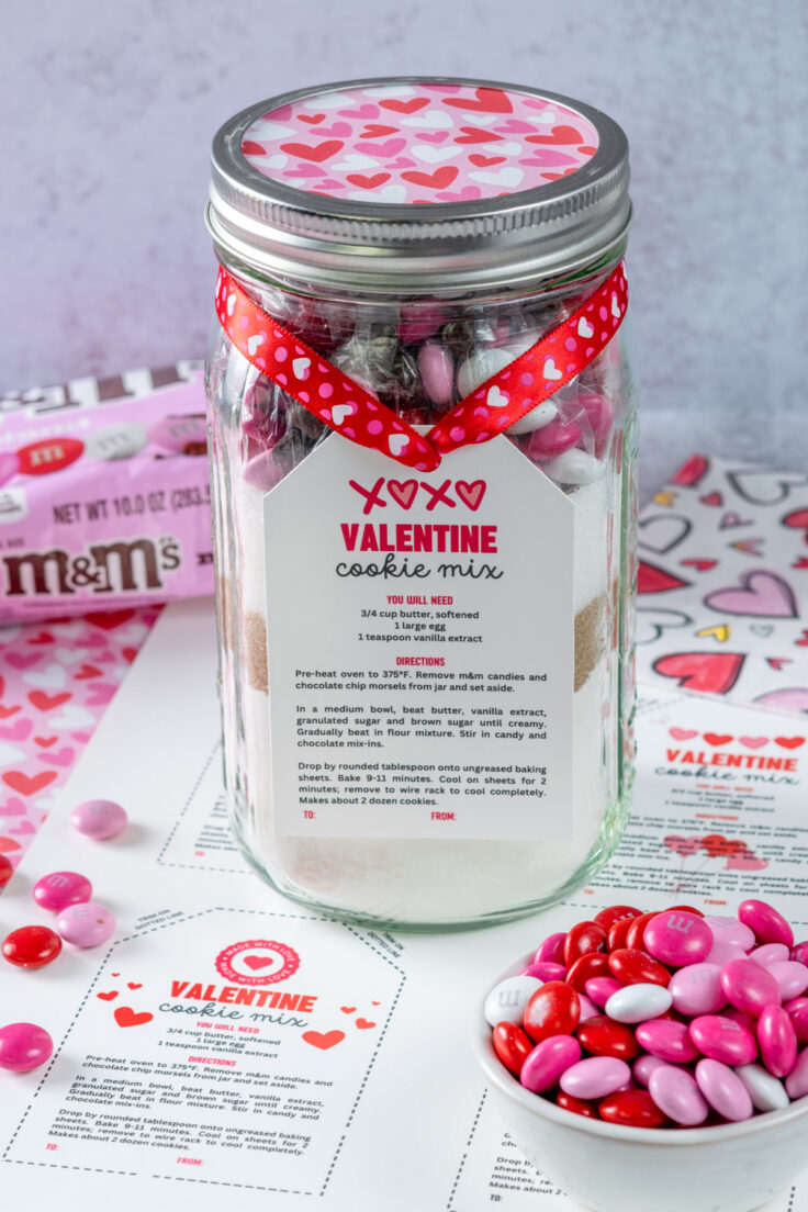 A jar of Valentine cookie mix sitting next to a bowl of pink M&M's, a bag of Cupid M&M's, and on top of a the gift tags that have been printed on paper.