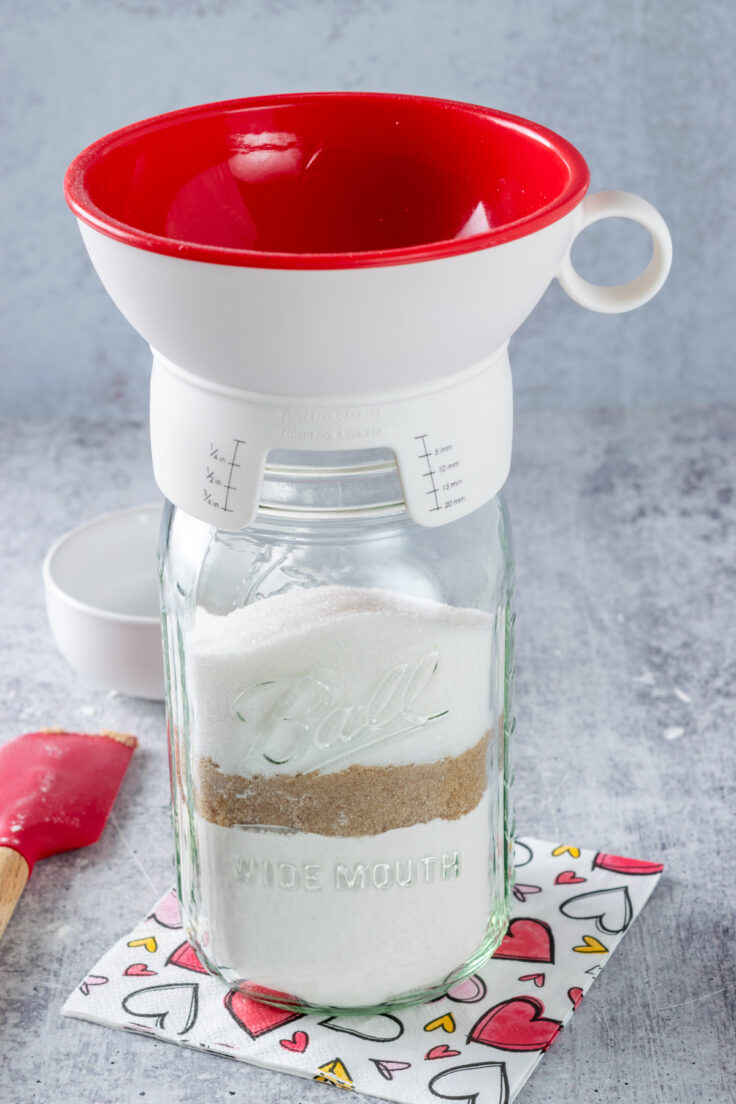 A canning funnel on a mason jar that has layers of flour, brown sugar and white sugar.