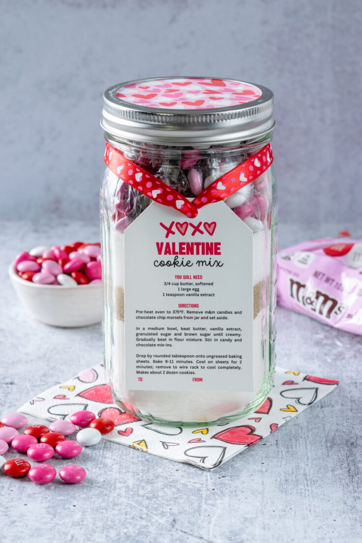 Mason jar Valentine Cookie mix in a jar that's decorated with ribbon and paper to give as a Valentine's Day gift. The jar is next to a small bowl of pink, white and red M&M's and the pink Cupid M&M's bag.