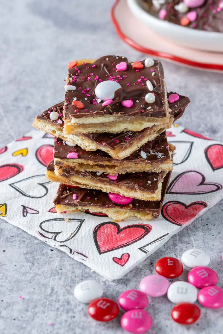 Saltine cracker toffee candy decorated with Valentine's Day sprinkles that sitting on a napkin with hearts all over it and next to some pink, red and white M&Ms candies strewn about.