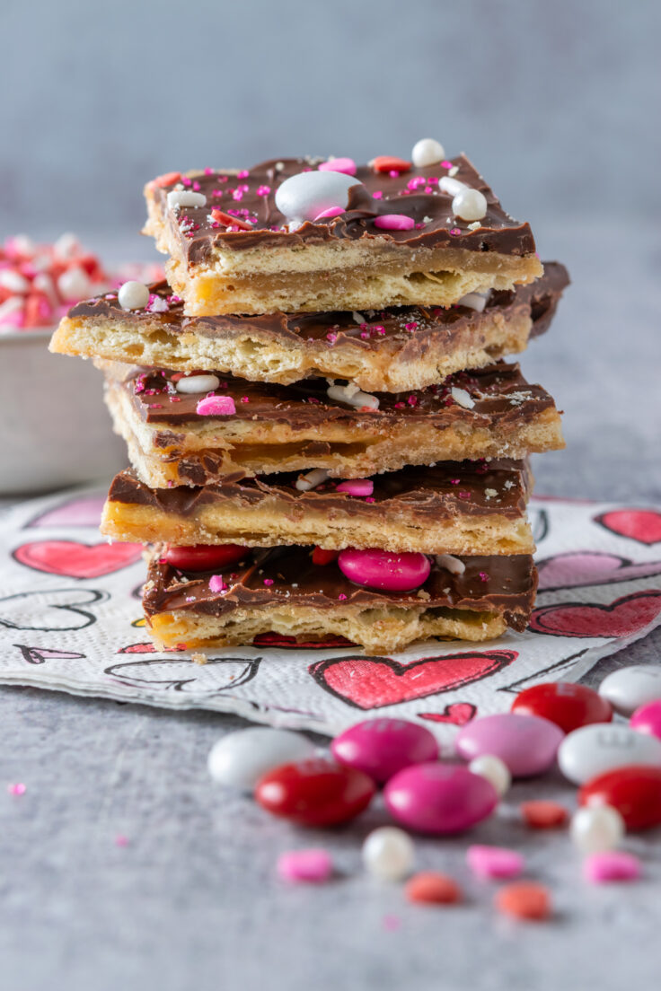 Close up view of a stack of saltine cracker candy sitting on a Valentine's Day napkin with hearts all over it. The side view of the candy shows the layers of crackers, toffee, chocolate and valentine sprinkles. There are some pink, red and white Valentine's Day M&Ms next to the crack candy and a small bowl full of Valentine's Day sprinkles.