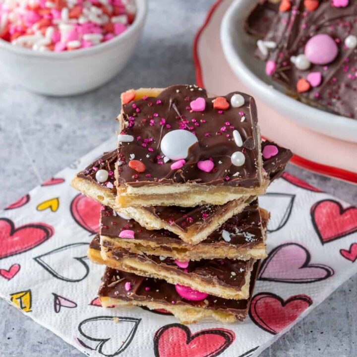 A stack of saltine cracker toffee candy decorated for Valentine's Day next to a plate with more crack candy and a small bowl full of pink, red and white Valentine sprinkles.