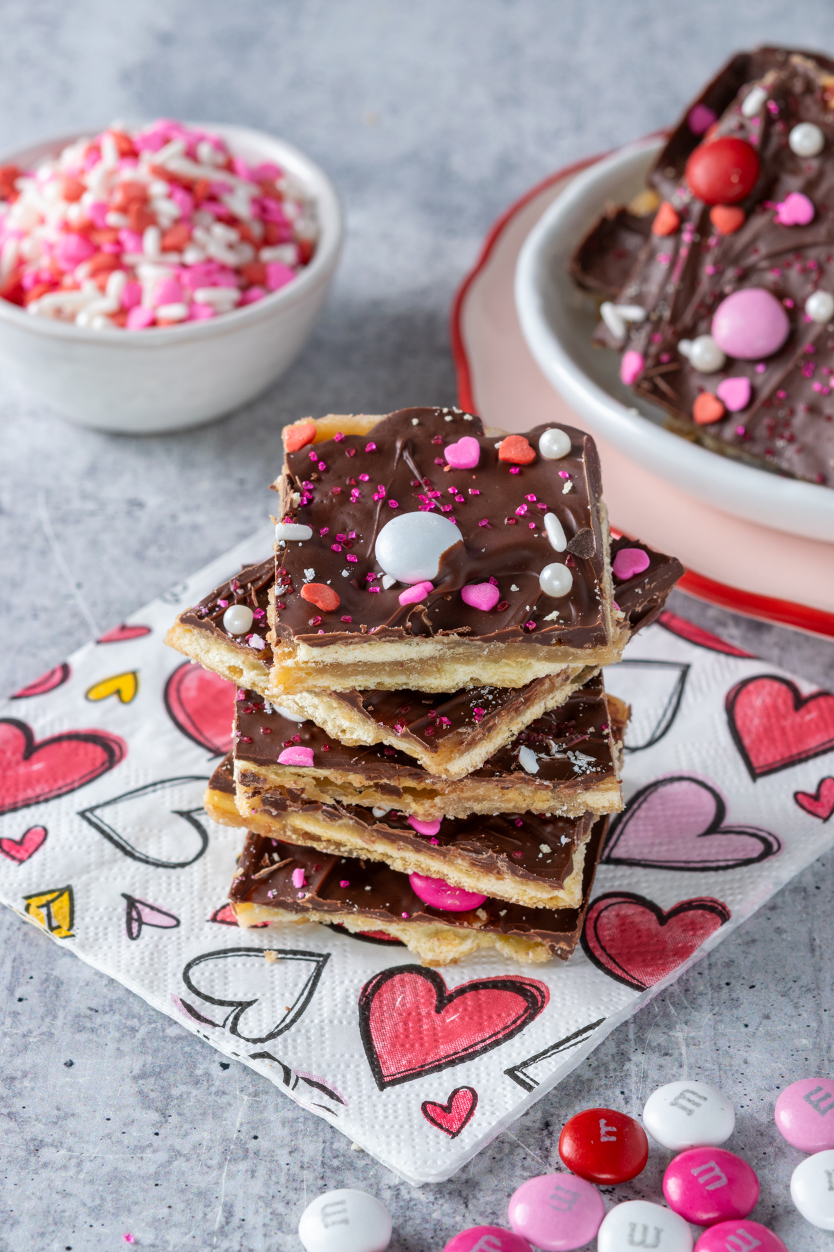 A stack of saltine cracker toffee candy decorated for Valentine's Day next to a plate with more crack candy and a small bowl full of pink, red and white Valentine sprinkles.