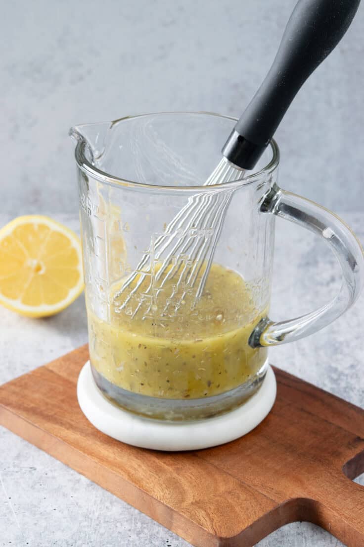 Homemade lemon vinaigrette salad dressing being whisked in a large glass measuring cup, with half of a cut lemon next to it.