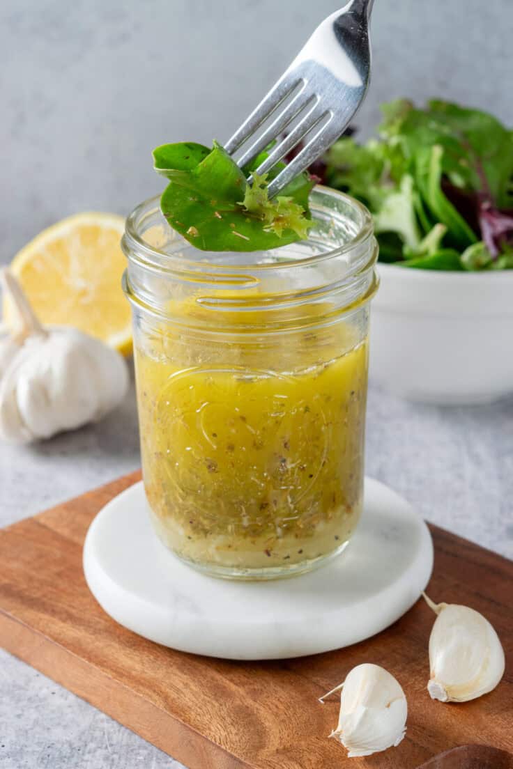 A fork of salad greens dripping in lemon salad dressing, that's held over the open jar of homemade dressing. There's half a lemon and a bowl of salad next to the jar of dressing.