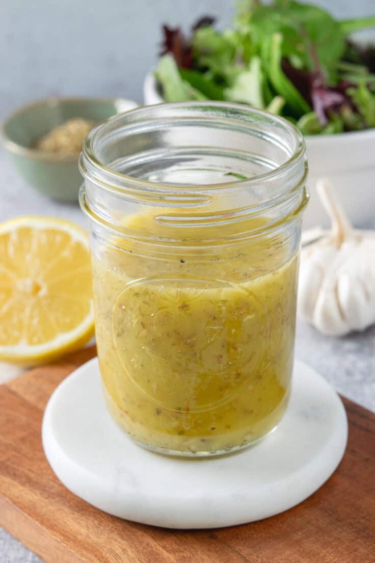 A small open mason jar filled with homemade vinaigrette. The jar is next to a lemon that's cut in half and a bulb of garlic.