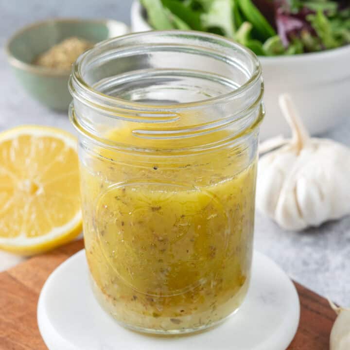 Homemade lemon salad dressing in a small glass jar, next to half a lemon, a bulb of garlic, and a bowl of salad greens.