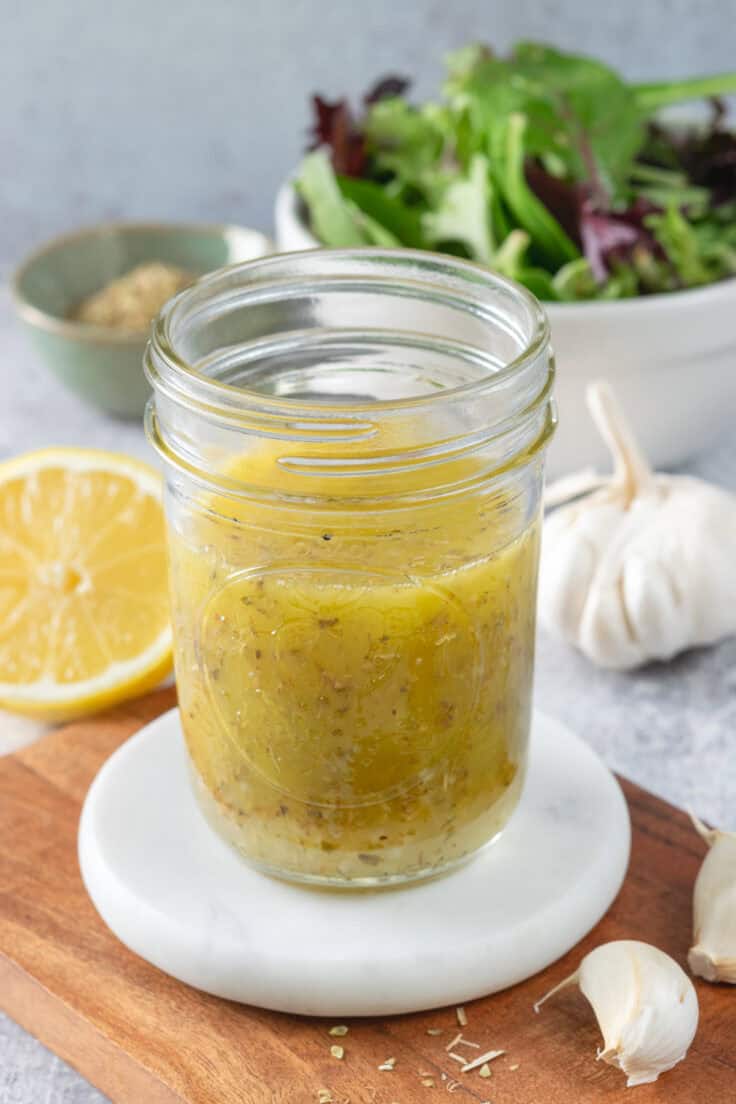 Homemade lemon salad dressing in a small glass jar, next to half a lemon, a bulb of garlic, and a bowl of salad greens.