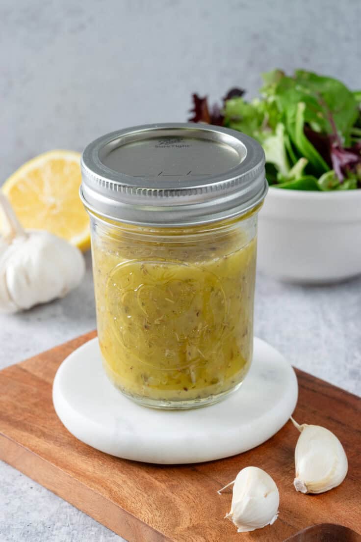 A mason jar filled with homemade lemon salad dressing. The jar of dressing is next to half a lemon, a bulb of garlic and a bowl of salad.