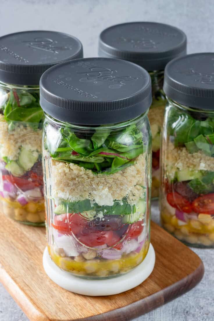 Four wide-mouth, quart-size mason jars meal prepped with layers of Mediterranean salad, beginning with the dressing on the bottom and salad greens on top.