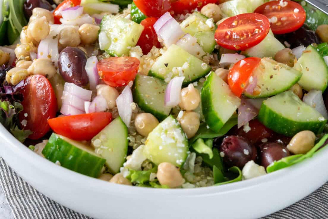 Close up view showing Mediterranean salad in a bowl, full of chopped onion, cucumber and tomatoes, chickpeas, quinoa, olives, feta cheese and salad greens, all coated in a lemon olive oil vinaigrette dressing.