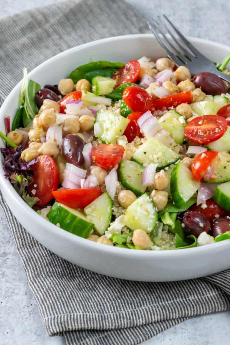 Mediterranean salad piled on a large plate with a fork, showing the cucumbers, tomatoes, olives, chickpeas, feta cheese and quinoa coated in lemon olive oil dressing, all on top of a bed of lettuce and salad greens.