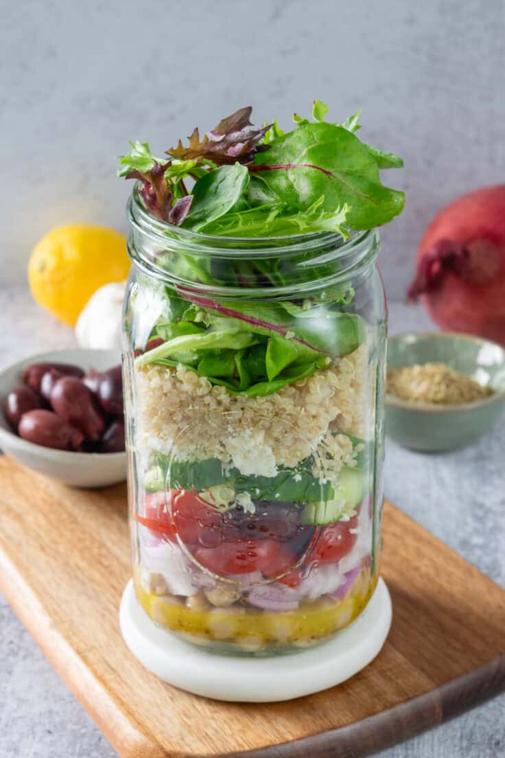 Mediterranean salad layered in a mason jar that's open with the salad greens popping out of the top. The salad in a jar is next to a bowl of Kalamata olives, oregano, garlic bulb, lemon and red onion.