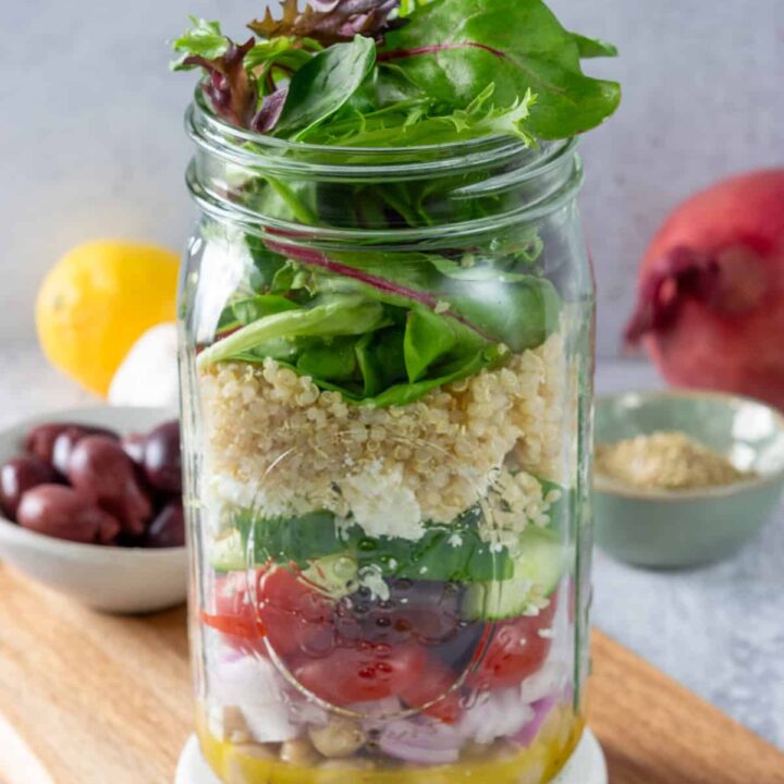 A Mediterranean salad packed in a mason jar with layers of lemon dressing, onion, tomatoes, cucumber, olives. feta cheese, quinoa and salad greens.