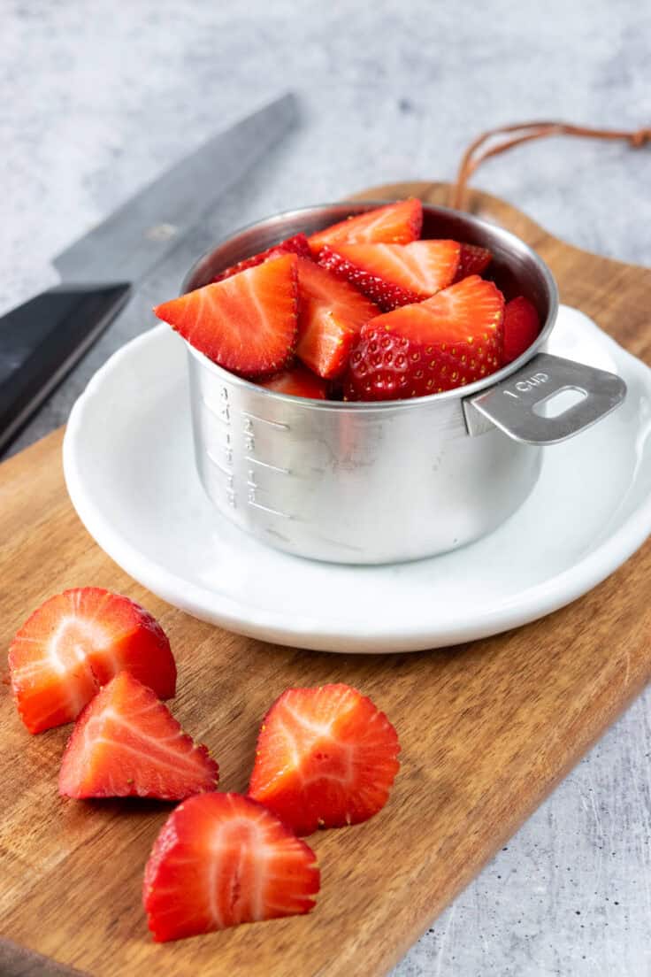 Sliced strawberries in a measuring cup which is on a small white plate. The strawberries are on a wooden cutting board and there is a knife in the background.