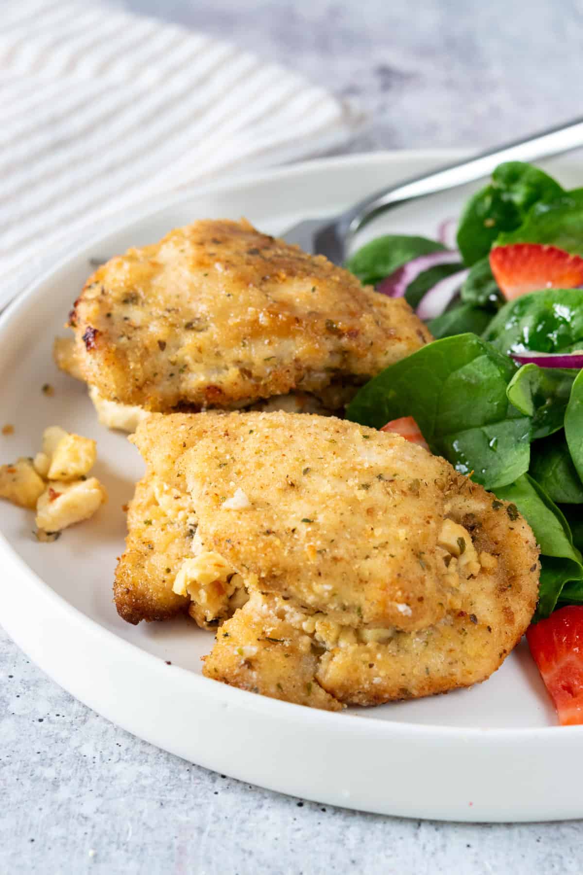 Two feta-stuffed chicken breasts on a dinner plate with a spinach salad next to it. There is a fork resting on the back of the plate and a tan striped napkin next to it.