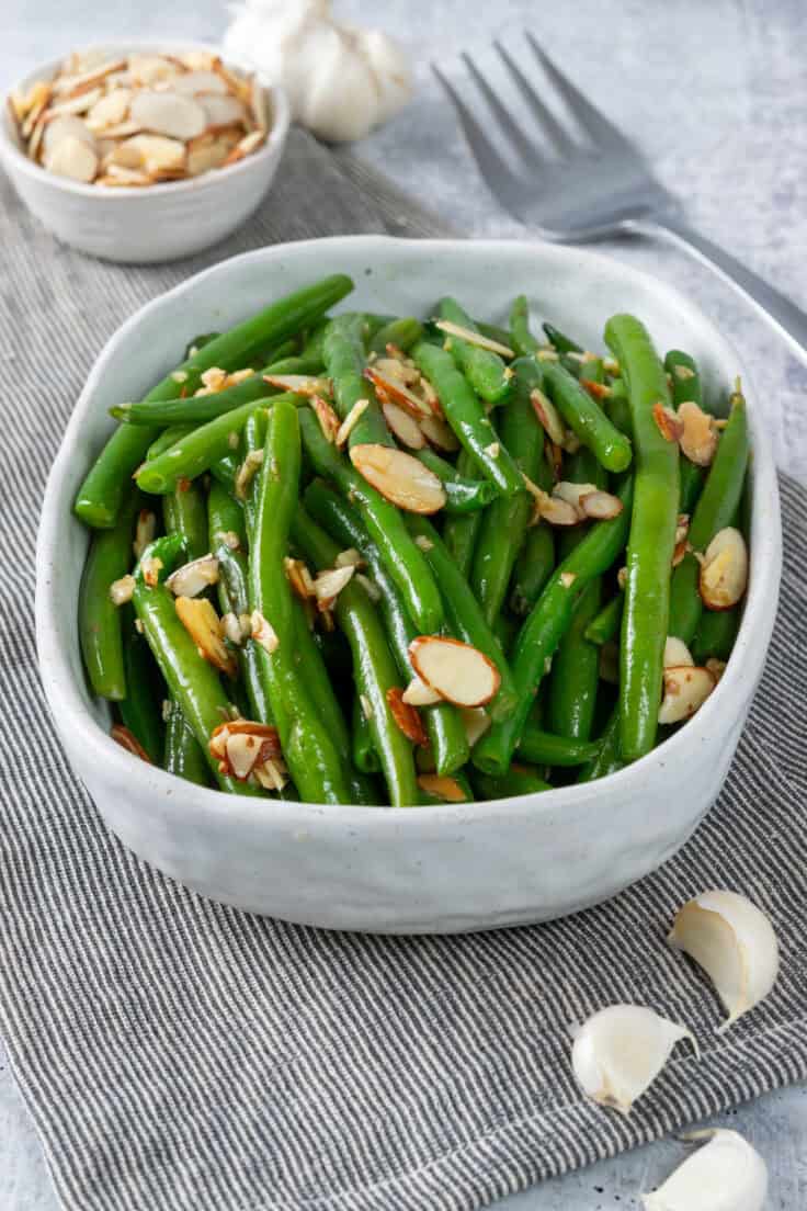 A serving dish with sauteed green beans with garlic and almonds. There's a small bowl of slivered almonds in the background and a few cloves of garlic in the foreground. To the right of the dish is a large serving fork.