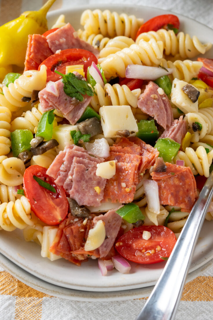 A close-up view of Italian pasta salad on a plate with a fork on it. The photo shows rotini noodles, salami, pepperoni, provolone cheese, olives, pepperoncini, tomatoes, green pepper, red oinion and parsley, covered in homemade Italian vinaigrette.