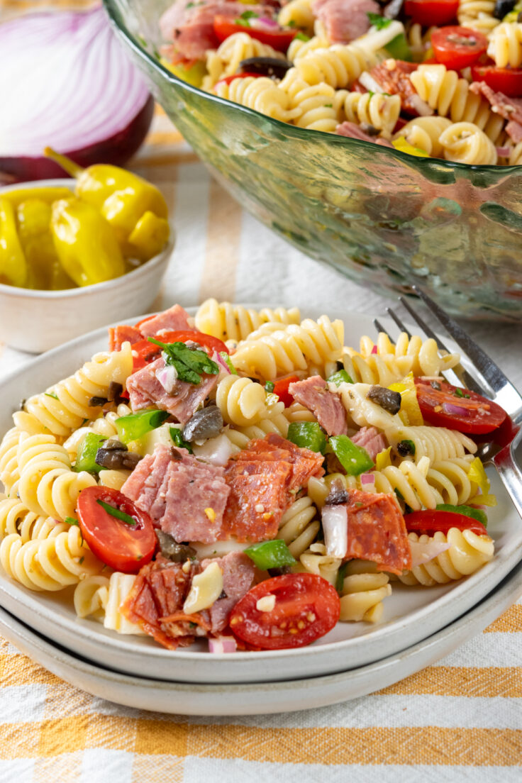 A plate of Italian pasta salad sitting on a table with a checkered table cloth. The plate is in front of a big bowl of the salad and a bowl of pepperoncini and half a red onion.
