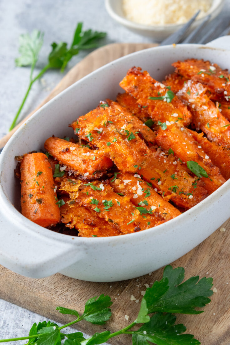 Parmesan roasted carrots in a small serving dish that's on a wooden board with some parsley leaves around it and a small bowl of grated parmesan cheese. The roasted carrots are garnished with finely chopped parsley.