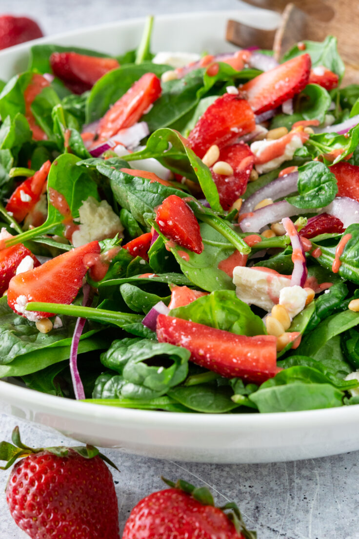 A close up view of a big bowl of strawberry spinach salad, showing cut up strawberries and pine nuts tossed with spinach, red onion and feta cheese.