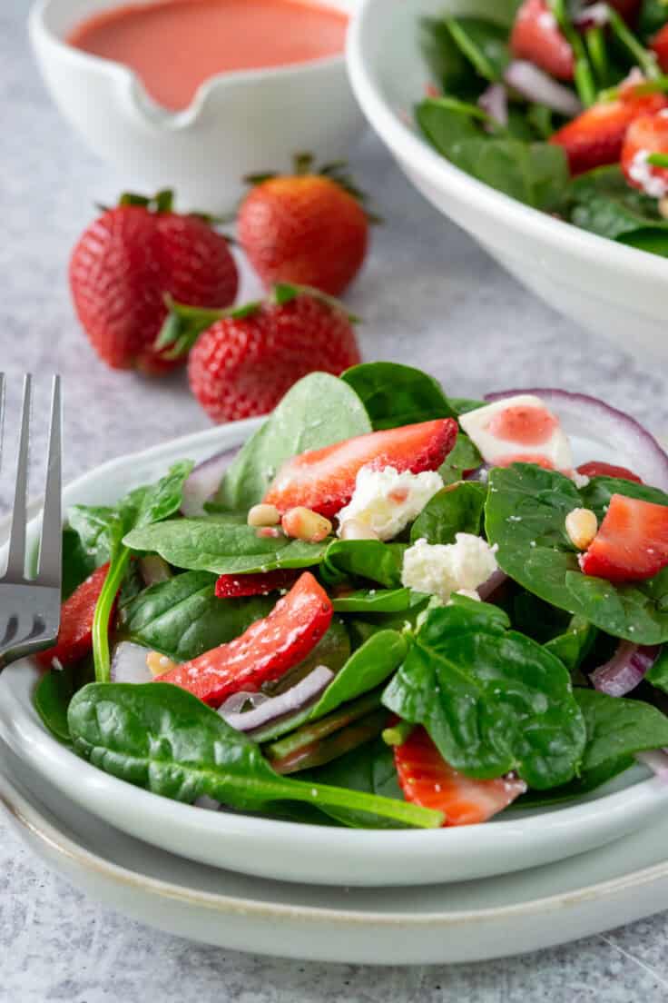 A side salad plate with spinach salad that has strawberries, red onion, feta cheese pine nuts and balsamic vinaigrette on it. Behind the side salad is three whole stawberries, a small pitcher of homemade vinaigrette and a large bowl of the salad.