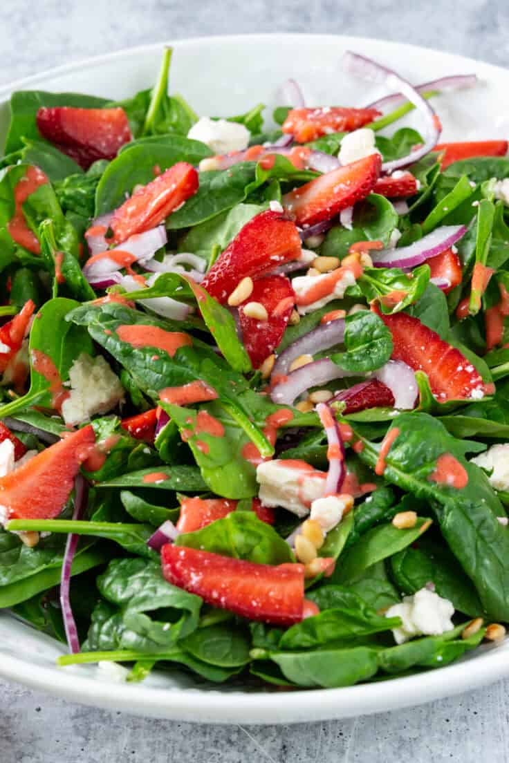 A large serving bowl of strawberry spinach salad that has homemade strawberry vinaigrette drizzled on top.