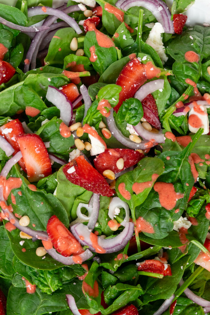 Close up view of a spinach salad with strawberries in it. You can see the strawberry vinaigrette dripping over the spinach, onion, feta, pine nuts and strawberries, showing how crisp and juicy the salad is.