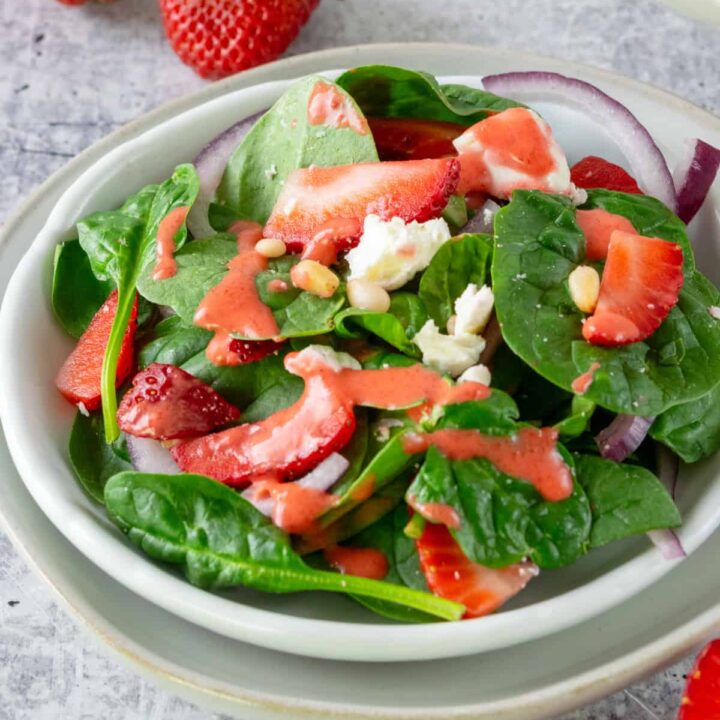 A spinach salad with strawberries and strawberry vinaigrette on a salad plate, which is next to a large bowl full of the strawberry spinach salad.