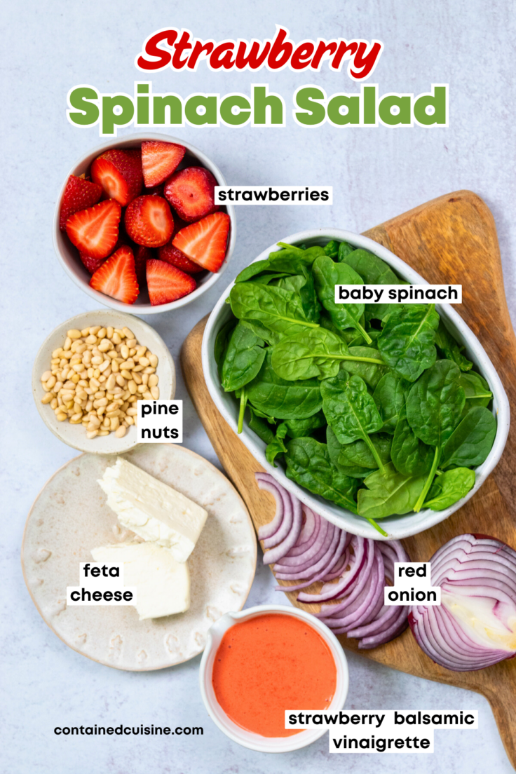 An overhead photo showing each ingredient needed to make this spinach salad, including a bowl of strawberries, bowl of baby spinach leaves, small bowl of pine nuts, block of feta cheese broken in half, sliced red onion, and a small bowl of homemade strawberry vinaigrette.