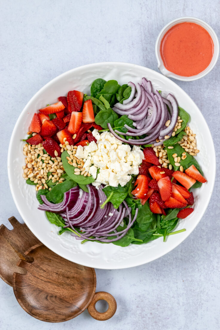 Overhead picture showing a large salad bowl with spinach in it. On top of the spinach is sections of sliced red onion, chopped strawberries, toasted pine nuts and crumbled feta cheese in the center. Next to the bowl are wooden salad forks for serving and a bowl of homemade strawberry vinaigrette.