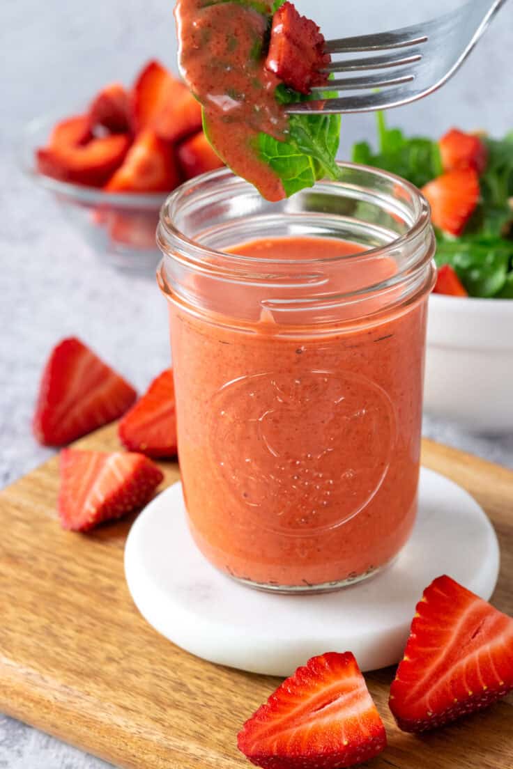 A fork with spinach leaves and a strawberry being dipped into jar of strawberry vinaigrette. The glass mason jar of dressing is on a white marble coaster which is on a wooden cutting board. There are cut strawberries around the vinaigrette and in a bowl in the background.