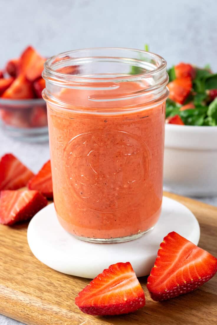 A pint mason jar full of pink-colored strawberry vinaigrette. In the background is a bowl of sliced strawberries and a small white bowl with a strawberry spinach salad in it.