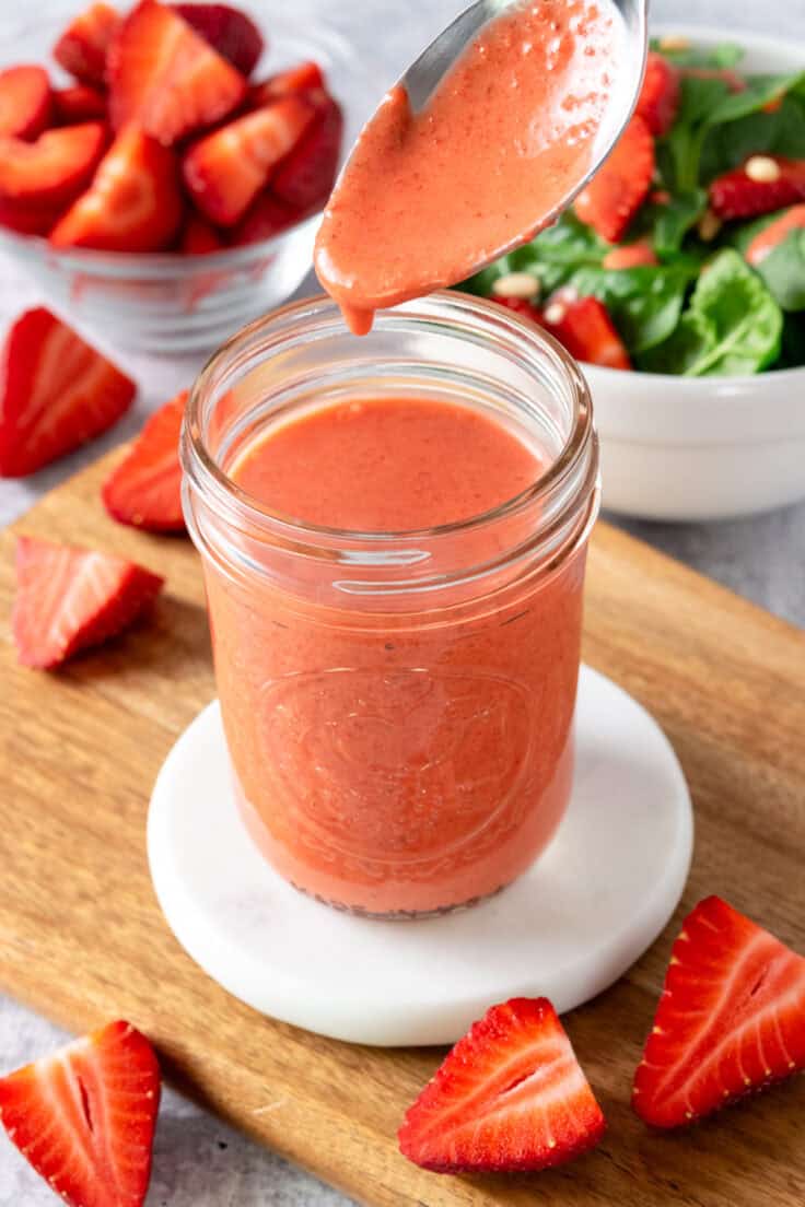 A spoon that's been dipped in the jar of homemade vinaigrette that's beneath it. In the background is a small glass bowl full of chopped strawberries and and another small bowl with spinach salad in it.