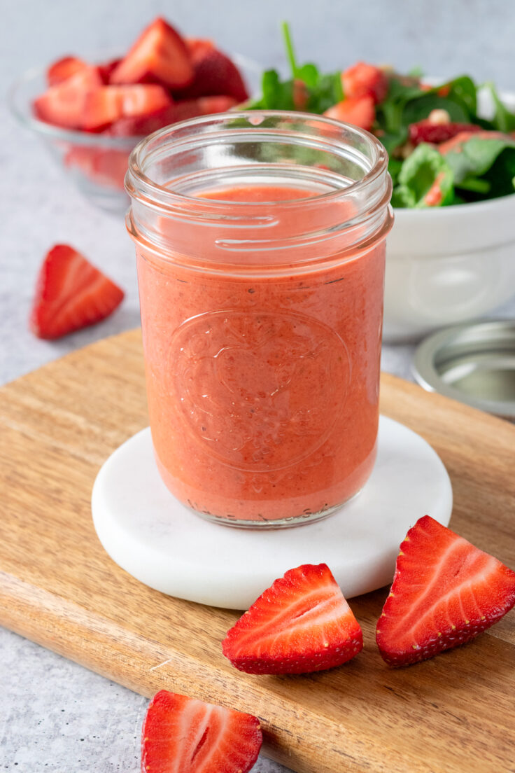 A small mason jar that contains homemade strawberry vinaigrette, which is in front of a bowl of spinach strawberry salad and a small glass bowl that has cut up strawberries in it.