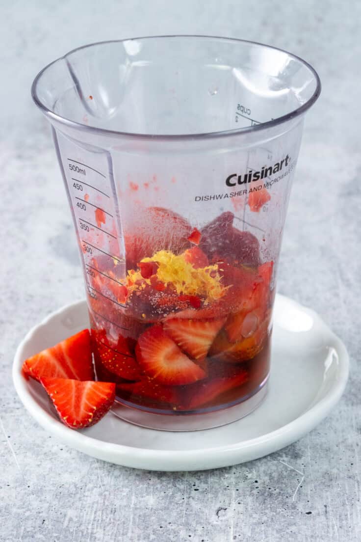 A tall measuring cup that has olive oil, vinegar, strawberries and lemon zest in it.